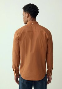 Pruun pikas varrukas särk, mis on valmistatud siledast kangast, nööpidega mansettide ja ümmarguse äärisega, nähtav tagant.