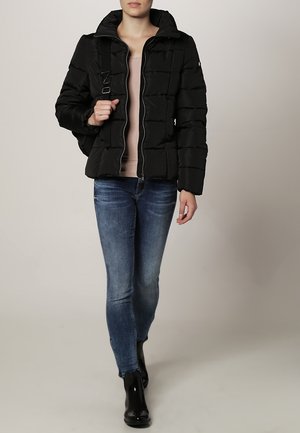 Kobieta idąca pieszo, ubrana w czarną kurtkę puchową, jasnoróżową bluzkę, obcisłe niebieskie dżinsy, czarne botki oraz niosąca na ramieniu czarną torbę.