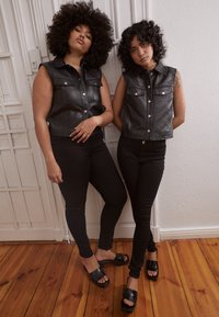 Duas modelos usam coletes sem mangas em imitação de pele preta com bolsos frontais e calças skinny pretas a condizer, combinados com sandálias escorregadias pretas brilhantes.