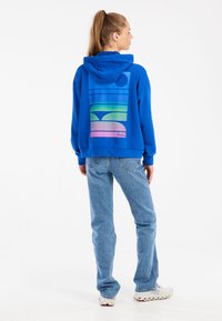 Sudadera azul con un gran diseño geométrico colorido en tonos degradados en la parte posterior. Combinada con jeans azul claro y zapatillas.