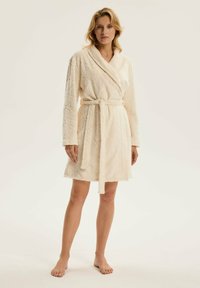 Knee-length, jasnobeżowy szlafrok wykonany z miękkiej, pikowanej tkaniny, z kołnierzem typu szal i pasującym paskiem. Ma długie rękawy i fakturowany wzór.