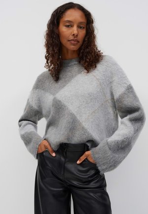 Woman with curly hair wearing a soft grey patterned sweater and high-waisted black leather pants with hands in pockets.