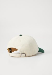 Gorra de béisbol hecha de lona crema con visera verde. Cuenta con una correa ajustable de metal y un botón verde en la parte superior.