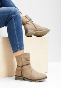 Stivaletti beige alla caviglia in un morbido materiale simile alla pelle scamosciata, progettati con una cerniera laterale e un nastro decorativo, con una suola in gomma bassa e texturizzata.