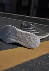 Chaussure de sport grise avec une tige texturée, présentant trois rayures blanches et une semelle en caoutchouc. La semelle a un relief à motifs pour une meilleure adhérence.