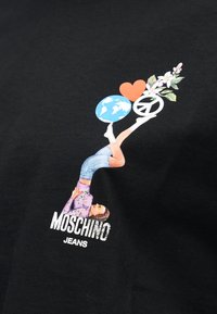 Άτομο ξαπλωμένο ανάσκελα με τα πόδια σηκωμένα, κρατώντας τη Γη, μια καρδιά, σύμβολο της ειρήνης και λουλούδια από πάνω, με το κείμενο "Moschino Jeans" από κάτω.