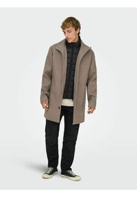 Cappotto in misto lana marrone con silhouette strutturata, chiusura con bottoni, collo alto, indossato sopra una giacca imbottita nera, abbinato a pantaloni neri e sneaker.