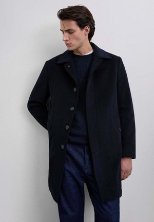 Abrigo de lana azul marino con cuello, cierre de cinco botones y bolsillos laterales, combinado con un jersey azul marino y jeans de denim. Textura suave, diseño minimalista.