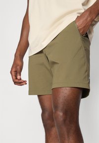 Pantaloni de plajă verde măslinie, confecționați dintr-un material ușor, dotati cu un buzunar cu fermoar și o margine finisată curat. Branding subtil aproape de margine.