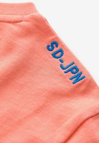 Korallfärgad bomullströja med blå broderad text "SD-JPN" på högra axeln. Slät textur med ribbat krage.
