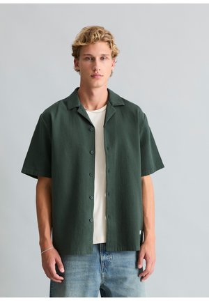 Jeune homme aux cheveux blonds courts et bouclés porte une chemise à manches courtes vert foncé boutonnée sur un t-shirt blanc et un jean bleu clair, debout contre un mur gris.