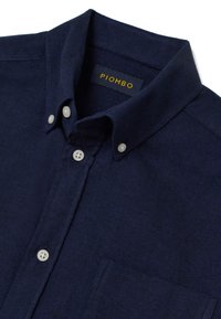 Camisa azul marino con patrón espiga, cuello abotonado, botones blancos y un bolsillo en el pecho. La etiqueta dice "PIOMBO." Textura lisa.