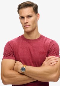 Camiseta roja jaspeada con cuello redondo, que presenta un pequeño logo bordado. Llevada con un reloj de pulsera de cuero marrón con esfera azul.