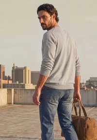 Homme aux cheveux foncés et à la barbe portant un pull gris et un jean, tenant un sac en cuir marron sur un toit avec des immeubles en arrière-plan.