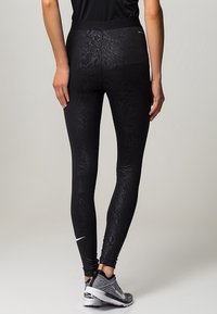 Leggings neri con un design testurizzato a stampa pitone, vita alta e un piccolo logo bianco. Indossati con scarpe da ginnastica grigie.