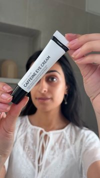 Frau hält eine Tube der The Inkey List Koffein-Augencreme vor ihr Gesicht und trägt eine weiße Bluse mit Spitzendetails.