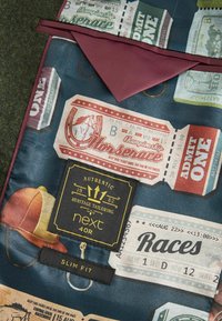 Tissu à motifs affichant des billets dans des teintes de marron, rouge et vert. Présente un col bordeaux et des étiquettes pour "prochain" et "ajusté".
