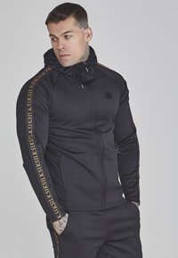 Schwarze Zip-Hoodie mit Kapuze und Kordelzug, ausgestattet mit goldenen Logo-Akzenten entlang der Ärmel. Hergestellt aus einem glatten, stretchigen Stoff.