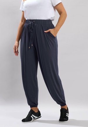 Vrouw draagt marineblauwe joggerbroek met elastische taille en trekkoord, zijzakken, elastische boorden, wit T-shirt en zwarte sneakers, staand voor een grijze achtergrond.