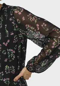 Abito nero trasparente con stampa floreale in rosa e verde. Maniche lunghe a sbuffo con polsini arricciati e scollatura con bottoni. Texture delicata.
