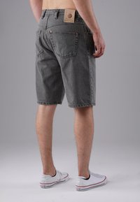 Man wearing gray denim shorts and white Converse sneakers, standing against a plain gray background, viewed from behind.