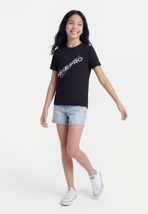 Fille aux longs cheveux noirs portant un t-shirt noir Nike Pro, un short en jean clair et des baskets blanches, souriante et posant sur un fond blanc.