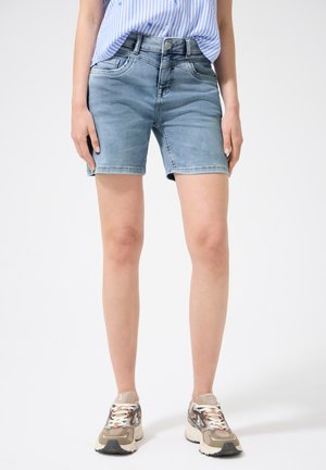 Personne portant un short en jean bleu clair et des baskets beiges, debout devant un fond blanc uni.