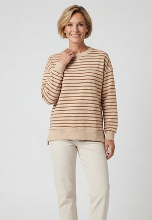 Lächelnde Frau, die ein beige-braun gestreiftes langärmliges Shirt und helle Hosen trägt und vor einem schlichten hellen Hintergrund steht.