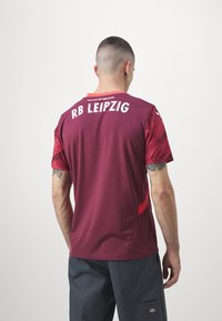 Maglietta sportiva color bordeaux con maniche rosse a trama. Presenta "RB LEIPZIG" in bianco sul retro e "PUOI FARE QUALSIASI COSA" sopra il colletto.