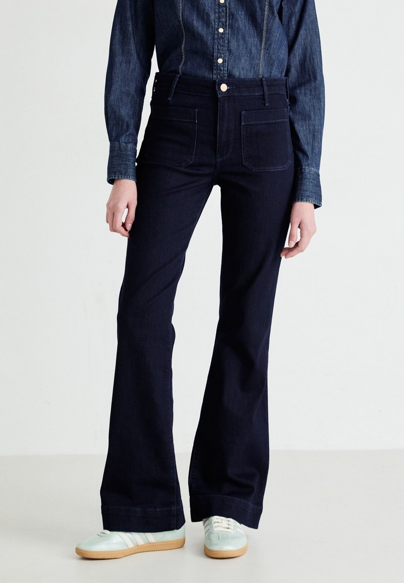 Mörk tvättade utsvängda jeans med hög midja, framfickor och kontrasterande sömmar, kombinerat med en denimskjorta med långa ärmar och ljusa sneakers.