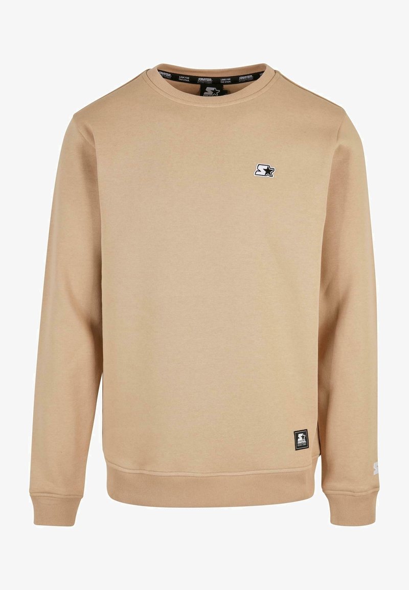 Sweat-shirt beige à manches longues avec col rond, petit logo étoile noir et blanc sur la poitrine et une étiquette de marque près de l'ourlet.