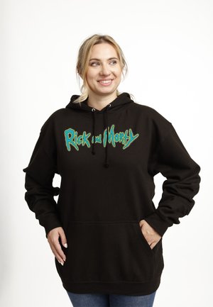 Smilende kvinde i en sort "Rick and Morty" hættetrøje med hænderne i forlommen, stående foran en ensfarvet hvid baggrund.