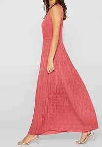 Růžová maxi šaty s plisovanou texturou, bez rukávů a s přiléhavým pasem. Spárovány se stříbrnými sandály na tenkých páscích.