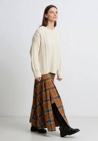 Kremowy sweter z grubym splotem, brązowa długa spódnica w kratę z bocznymi rozcięciami, czarne kozaki za kolano z klamrami. Prosta, teksturowana i warstwowa stylizacja.