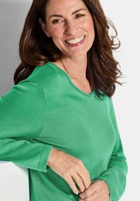 Frau mit braunen Haaren, die lächelt, trägt ein grünes langärmliges Shirt und lehnt an einer weißen Oberfläche vor einem hellen Hintergrund.