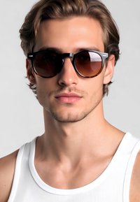 Gli occhiali da sole tondeggianti presentano una montatura in tartaruga e lenti marroni scure. Il modello indossa una canottiera bianca e ha capelli ondulati di lunghezza media.