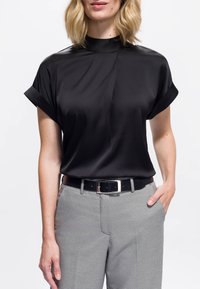Mujer con una blusa negra de manga corta y cuello alto metida dentro de pantalones grises de estampado, llevando un cinturón negro, con una mano en el bolsillo.