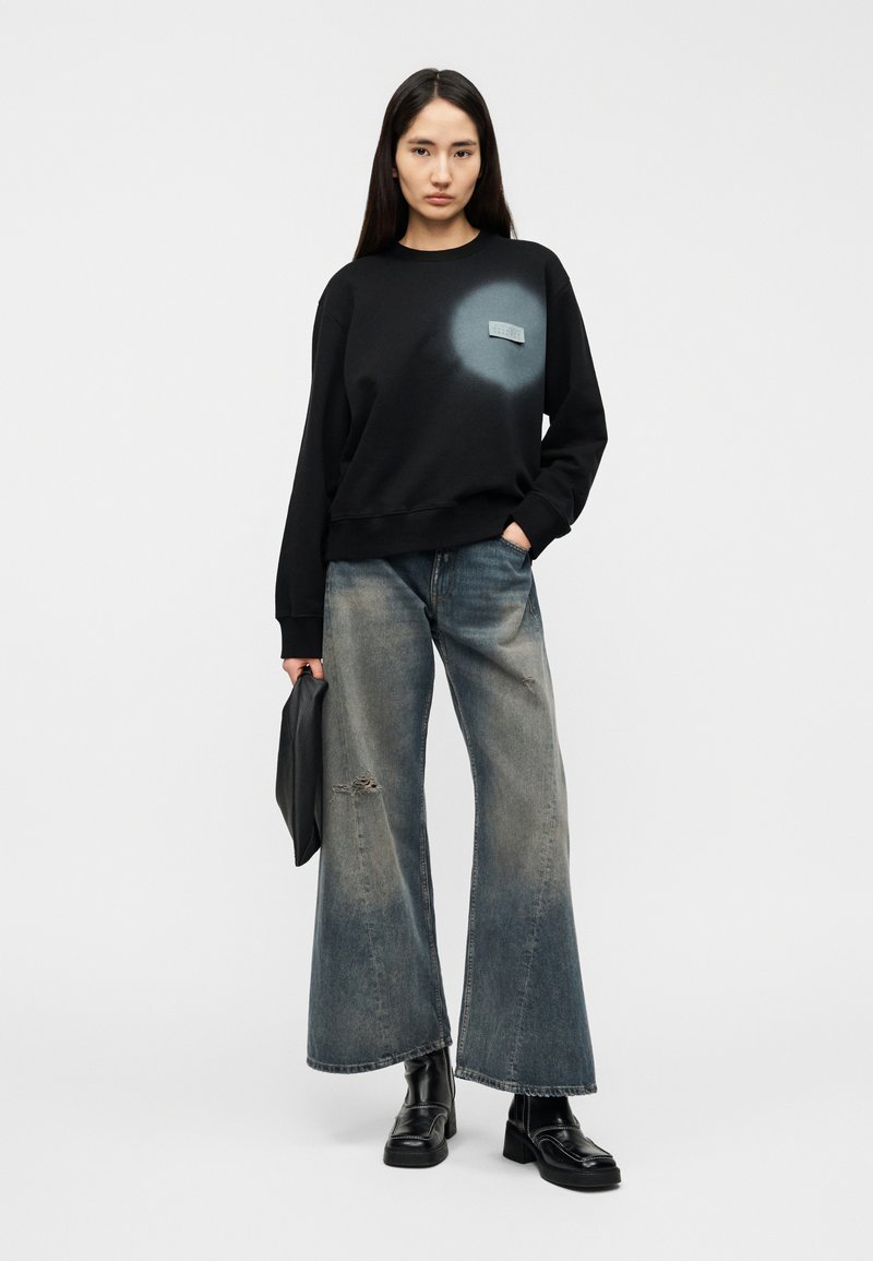 Sweat-shirt noir avec un graphique bleu délavé, associé à un jean à jambes larges et délavé clair, avec des détails usés. Bottines noires et une pochette.