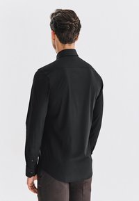 Czarna koszula z długim rękawem i wysokim kołnierzem, gładka w dotyku, z przeszyciem na karczku z tyłu i mankietami zapinanymi na guziki. Noszona z ciemnobrązowymi spodniami.