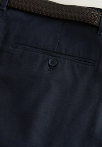 Tela azul marino de pantalones que presenta un cinturón marrón tejido, un bolsillo trasero con un botón negro y una superficie texturizada.