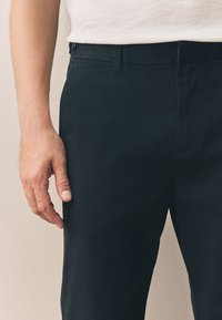 Pantalon en coton noir à la texture lisse, doté d'une poche avant et d'une coupe ajustée, agrémenté d'un détail de fermeture par bouton.