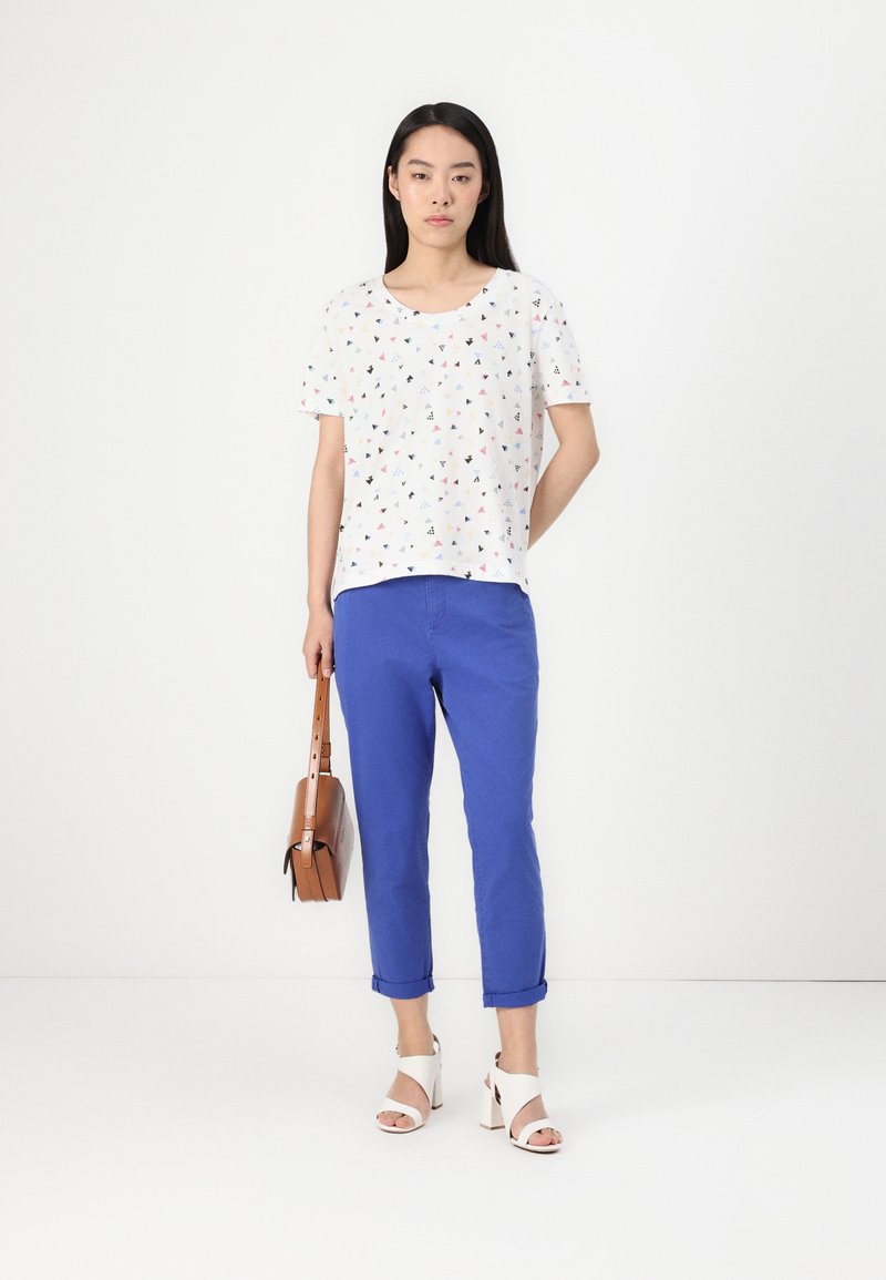 T-shirt blanc avec un imprimé triangulaire multicolore, associé à un pantalon bleu vif retroussé. Sac en bandoulière marron et sandales à talons blanches.