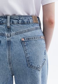 Vaaleansiniset farkut, joissa on korkea vyötärö, ruskea nahkalamina, kupariset niitit ja kaksi takataskua, joissa on näkyvät ompeleet.