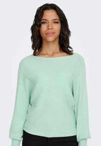 Maglione verde menta a coste con ampio scollo a barca e maniche lunghe, caratterizzato da un tessuto strutturato e una vestibilità rilassata. Abbinato a pantaloni neri.
