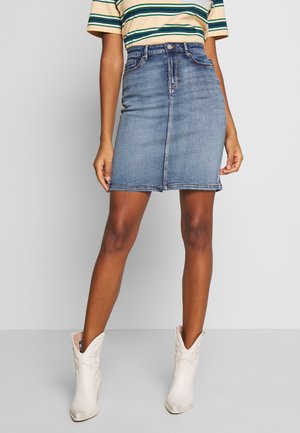 Femme portant une jupe en jean bleue, un t-shirt à manches courtes rayé et des bottines blanches, debout devant un fond uni.