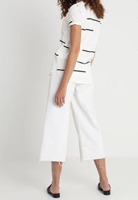 Vit kortärmad skjorta med svarta horisontella ränder, i kombination med vida vita byxor med fransade fållar och svarta slip-on skor.