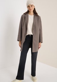 Manteau en laine brun clair avec des revers, fermeture à un bouton, associé à un t-shirt blanc et un jean sombre, complété par un bonnet en tricot.