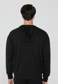 Felpa nera con cappuccio realizzata in tessuto morbido. Presenta una tasca a marsupio, polsini a coste e un cappuccio regolabile con cordino. Design semplice senza pattern visibili.