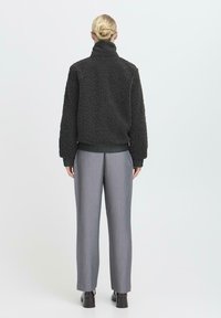 Czarna polarowa kurtka z wysokim kołnierzem, teksturowany materiał, ściągacze rękawów i dół, w zestawieniu z szarymi spodniami typu wide-leg. Gładkie, eleganckie obuwie.