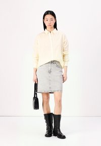 Gul stripe oversized skjorte, grå denim skjørt, svarte kne-høye støvler, holder en liten svart håndveske. Enfarget bakgrunn, minimale tilbehør.